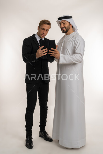 Hosting foreign businessmen in the United Arab Emirates, exchanging experiences and knowledge between countries, Portrait of an Arab Gulf Emirati man wearing a kandura and ghutra standing next to a young man wearing a formal suit working on a tablet, full body image, white background