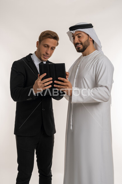 تبادل الخبرات والمعرفة بين البلاد, استضافة رجال الاعمال الاجانب في الامارات العربية المتحدة, بورتريه لرجل عربي خليجي اماراتي يرتدي الكندورة والغترة يقف بجانب شاب اجبني يرتدي البدلة الرسمية يقومان بالعمل على جهاز التابلت، خلفية بيضاء