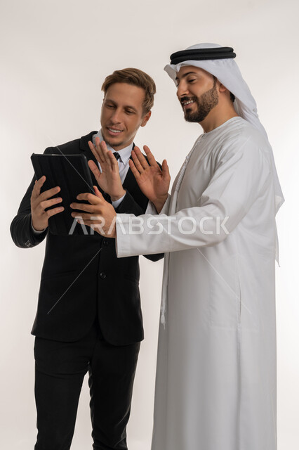 Exchange of expertise and knowledge between countries, hosting foreign businessmen in the United Arab Emirates, portrait of an Arab Gulf Emirati man wearing a kandura and ghutra standing next to a young man wearing a formal suit, working on a tablet, white background