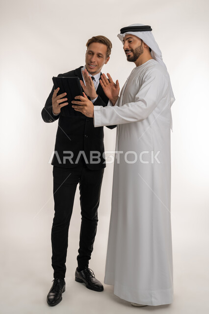 تعابير السرور والسعادة، استضافة رجال الاعمال الاجانب في الامارات العربية المتحدة, تبادل الخبرات والمعرفة بين البلاد, بورتريه لرجل عربي خليجي اماراتي يرتدي الكندورة والغترة يقف بجانب شاب اجبني يرتدي البدلة الرسمية يقومان بالعمل على التابلت، الجسم الكامل، خلفية بيضاء