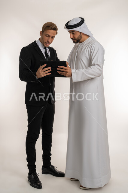 استضافة رجال الاعمال الاجانب في الامارات العربية المتحدة, تبادل الخبرات والمعرفة بين البلاد, بورتريه لرجل عربي خليجي اماراتي يرتدي الكندورة والغترة يقف بجانب شاب اجبني يرتدي البدلة الرسمية يقومان بالعمل على جهازالتابلت، صورة لكامل الجسم، خلفية بيضاء