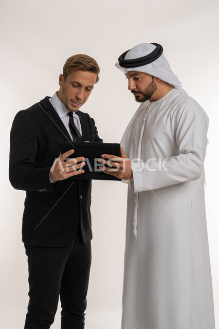 تبادل الخبرات والمعرفة بين البلاد, استضافة رجال الاعمال الاجانب في الامارات العربية المتحدة, بورتريه لرجل عربي خليجي اماراتي يرتدي الكندورة والغترة يقف بجانب شاب اجبني يرتدي البدلة الرسمية يقومان بالعمل على جهاز التابلت، خلفية بيضاء
