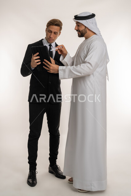 استضافة رجال الاعمال الاجانب في الامارات العربية المتحدة, تبادل الخبرات والمعرفة بين البلاد, بورتريه لرجل عربي خليجي اماراتي يرتدي الكندورة والغترة يقف بجانب شاب اجبني يرتدي البدلة الرسمية يقومان بالعمل على جهازالتابلت، صورة لكامل الجسم، خلفية بيضاء