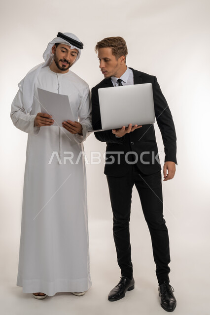 تبادل الخبرات والمعرفة بين البلاد, استضافة رجال الاعمال الاجانب في الامارات العربية المتحدة, بورتريه لرجل عربي خليجي اماراتي يرتدي الكندورة والغترة يقف بجانب شاب اجبني يرتدي البدلة الرسمية يقومان بالعمل على الحاسوب المحمول، صورة لكامل الجسم، خلفية بيضاء