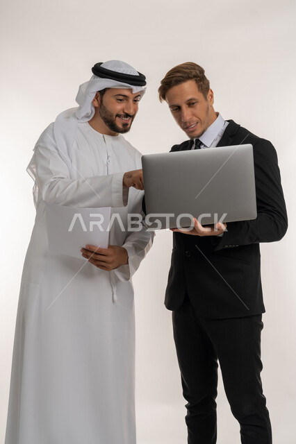 استضافة رجال الاعمال الاجانب في الامارات العربية المتحدة, تبادل الخبرات والمعرفة بين البلاد, بورتريه لرجل عربي خليجي اماراتي يرتدي الكندورة والغترة يقف بجانب شاب اجبني يرتدي البدلة الرسمية يقومان بالعمل على الحاسوب المحمول، خلفية بيضاء