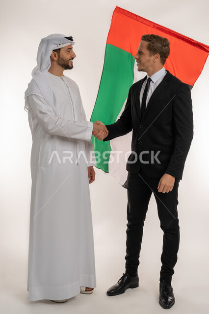 Raising the country's flag in celebration of the National Day on December 2, agreement and partnership on business and commercial projects, portrait of an Arab Gulf Emirati man wearing a kandura and ghutra shaking hands with a young foreigner wearing a formal suit, learning about the culture of the people of the Emirates, full body image, white background