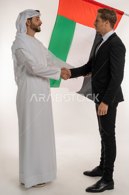 الإتفاق والشراكة على الأعمال والمشاريع التجارية، رفع علم البلاد احتفالا بالعيد الوطني 2 ديسمبر، بورتريه لرجل عربي خليجي اماراتي يرتدي الكندورة والغترة يقوم بمصافحة شاب اجنبي يرتدي البدلة الرسمية، التعرف على ثقافة شعب الامارات، صورة لكامل الجسم، خلفية بيضاء