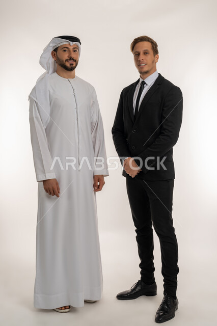 استضافة رجال الاعمال الاجانب في الامارات العربية المتحدة, تبادل الخبرات والمعرفة بين البلاد, بورتريه لرجل عربي خليجي اماراتي يرتدي الكندورة والغترة يقف بجانب سائح اجبني يرتدي البدلة الرسمية ينظران الى الكاميرا بتعابير السرور, صورة لكامل الجسم، خلفية بيضاء