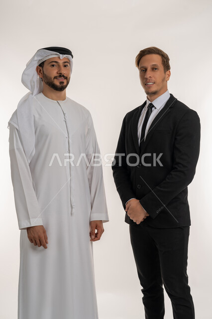 Exchange of expertise and knowledge between countries, hosting foreign businessmen in the United Arab Emirates, portrait of an Arab Gulf Emirati man wearing a kandura and ghutra standing next to a tourist, answer me, wearing a formal suit, looking at the camera with expressions of pleasure, white background
