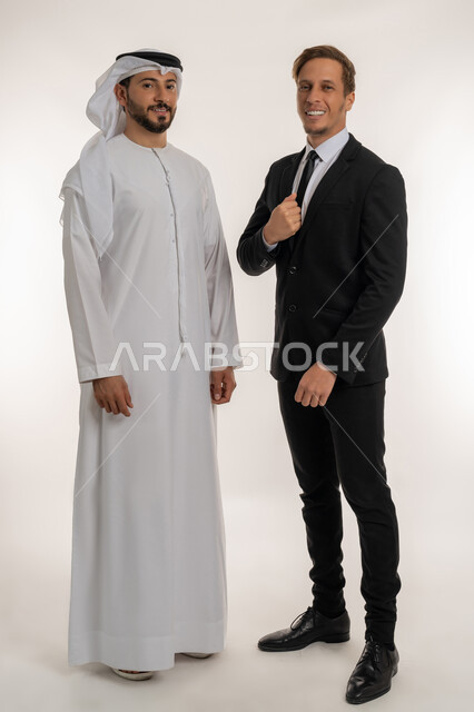 استضافة رجال الاعمال الاجانب في الامارات العربية المتحدة, تبادل الخبرات والمعرفة بين البلاد, بورتريه لرجل عربي خليجي اماراتي يرتدي الكندورة والغترة يقف بجانب سائح اجبني يرتدي البدلة الرسمية ينظران الى الكاميرا بتعابير السرور, صورة لكامل الجسم، خلفية بيضاء