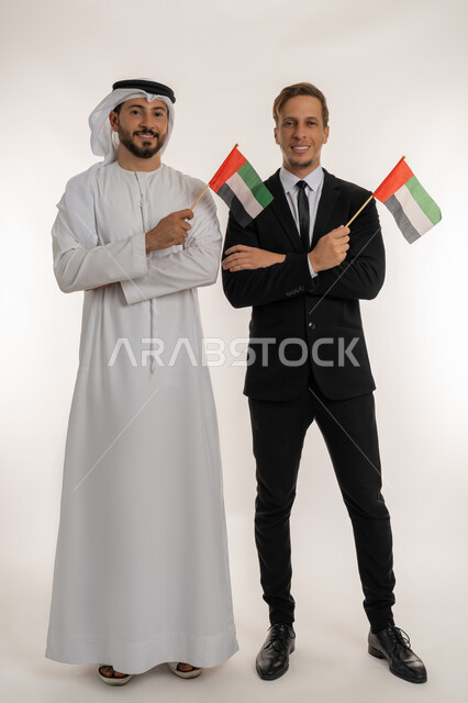 ذكرى تأسيس الاتحاد 1971، التعرف على تاريخ الامارات، بورتريه لشاب اجنبي يرتدي البدلة الرسمية يقف بجانب رجل اماراتي يرتدي الغترة والكندورة يحملان العلم للاحتفال باليوم بالوطني الاماراتي 2 ديسمبر، النظر الى الكاميرا بتعابير الفخر، الجسم بالطول الكامل، خلفية بيضاء