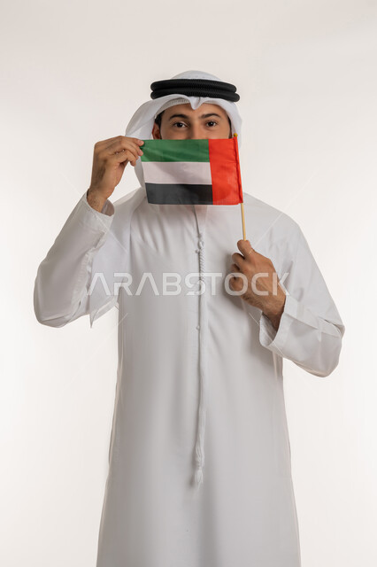 النظر إلى الكاميرا بإيماءات الاعتزاز والفخر، الاحتفال باليوم الوطني 2 ديسمبر، إحياء ذكرى استقلال البلاد1971، مفهوم حب الوطن والانتماء إليه، بورتريه لرجل عربي خليجي إماراتي يرتدي الكندورة والغترة يرفع علم الإمارات العربية المتحدة امام فمه، خلفية بيضاء