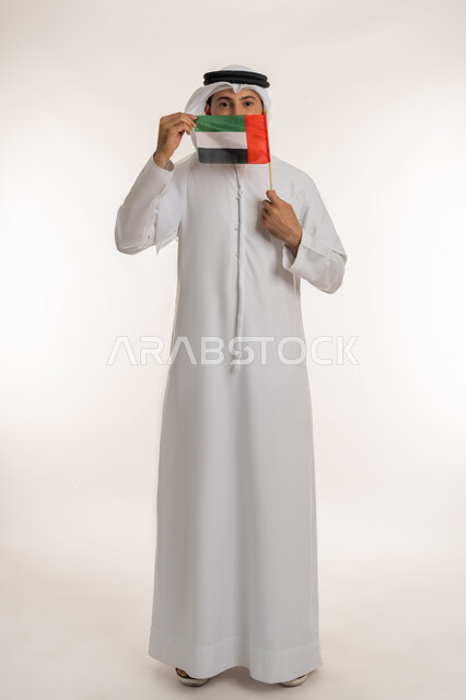 النظر إلى الكاميرا بإيماءات الاعتزاز، الاحتفال باليوم الوطني 2 ديسمبر، إحياء ذكرى استقلال البلاد1971، مفهوم حب الوطن والانتماء إليه، بورتريه لرجل عربي خليجي إماراتي يرتدي الكندورة والغترة يرفع علم الإمارات العربية المتحدة امام فمه، صورة لكامل الجسم، خلفية بيضاء