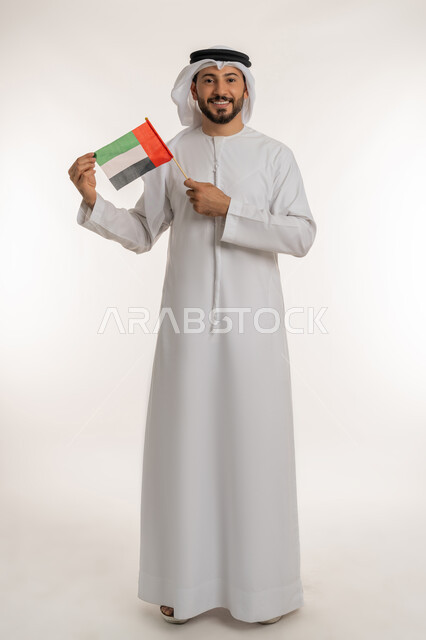 إحياء ذكرى استقلال البلاد1971، مفهوم حب الوطن والانتماء إليه، بورتريه لرجل عربي خليجي إماراتي يرتدي الكندورة والغترة يرفع علم الإمارات العربية المتحدة، الاحتفال باليوم الوطني 2 ديسمبر، النظر إلى الكاميرا بإيماءات الاعتزاز والفخر، صورة لكامل الجسم، خلفية بيضاء