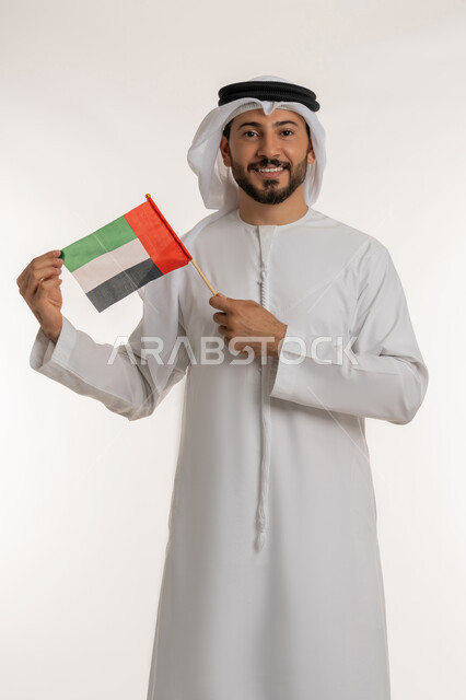 The concept of patriotism and belonging to the homeland, commemorating the country's independence in 1971, a portrait of an Emirati Gulf Arab man wearing a kandura and ghutra raising the flag of the United Arab Emirates, celebrating the National Day on December 2, looking at the camera with gestures of pride and honor, white background