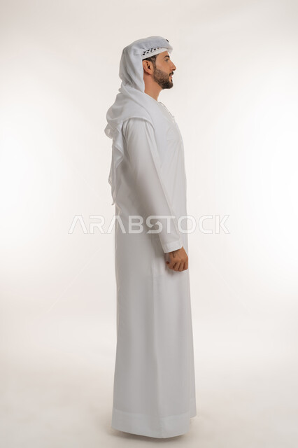 Standing straight and looking somewhere with gestures of self-confidence, expressions of seriousness and firmness, interest in outward appearance, concept of elegance, masculinity and strength, side portrait of an Arab Gulf Emirati man wearing an Emirati kandura and ghutra looking at something, full body image, white background