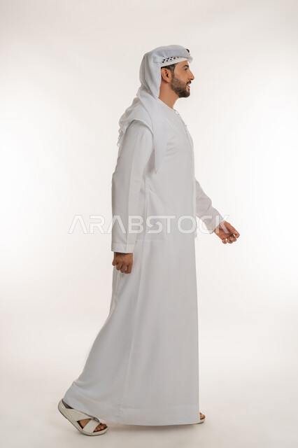 The concept of elegance, masculinity and strength, interest in outward appearance, moving forward with steady steps, gestures of walking and going to a certain place, a side portrait of an Arab Gulf Emirati man wearing a kandura and a ghutra looking at something with expressions of pleasure and happiness, a full body image, white background