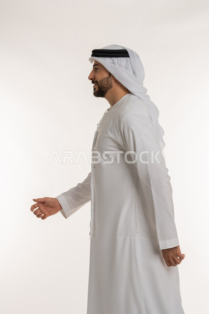 التقدم إلى الأمام بخطوات ثابتة، تعابير البشاشة والسرور، الاهتمام بالمظهر الخارجي، مفهوم الأناقة والرجولة والقوة, ايماءات المشي و الذهاب إلى مكان معين، بورتريه من الجانب لرجل عربي خليجي إماراتي يرتدي الكندورة والغترة ينظر إلى شيء ما، خلفية بيضاء