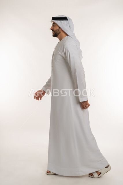 ايماءات المشي و الذهاب إلى مكان معين، بورتريه من الجانب لرجل عربي خليجي إماراتي يرتدي الكندورة والغترة ينظر إلى شيء ما، التقدم إلى الأمام بخطوات ثابتة، تعابير البشاشة والسرور، الاهتمام بالمظهر الخارجي، مفهوم الأناقة والرجولة , الطول الكامل للجسم، خلفية بيضاء