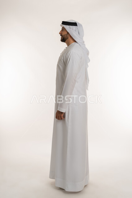 Standing straight and looking to the left with gestures of self-confidence, expressions of seriousness and firmness, interest in outward appearance, concept of elegance, masculinity and strength, side portrait of an Arab Gulf Emirati man wearing an Emirati kandura and ghutra looking at something, full body length, white background