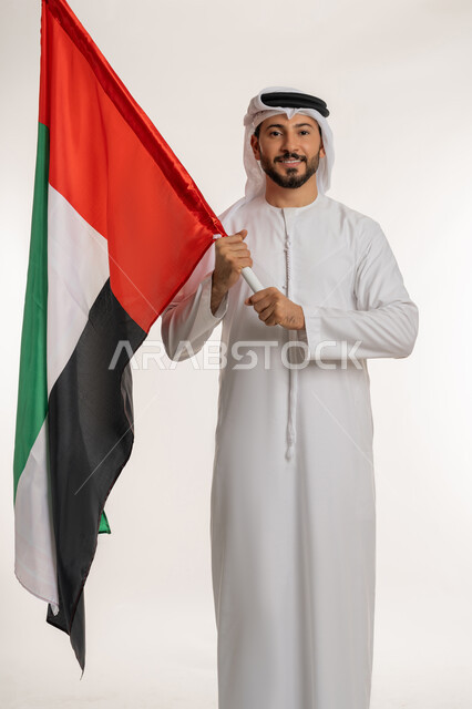الاحتفال باليوم الوطني الاماراتي 2 ديسمبر، يوم استقلال الإمارات 1971 ، الوقوف باستقامة والنظر إلى الكاميرا بتعابير الفخر، بورتريه لرجل عربي خليجي إماراتي يرتدي الكندورة والغترة يحمل بيده علم الإمارات العربية المتحدة، الطول الكامل للجسم، خلفية بيضاء