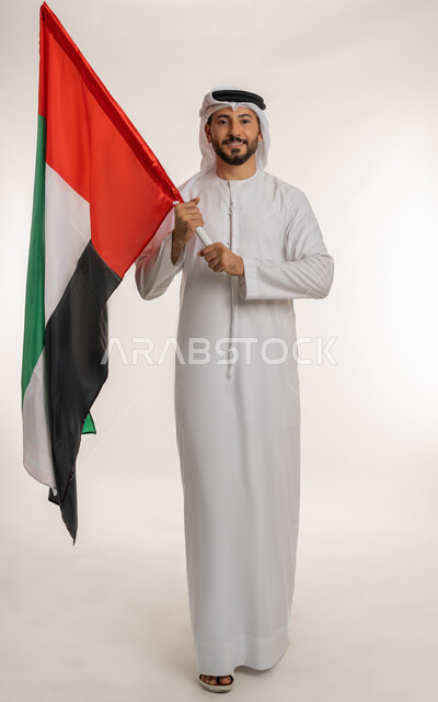 الوقوف باستقامة والنظر إلى الكاميرا بتعابير الفخر، بورتريه لرجل عربي خليجي إماراتي يرتدي الكندورة والغترة يحمل بيده علم الإمارات العربية المتحدة، هوية اليوم الوطني الاماراتي 2 ديسمبر، يوم استقلال الإمارات 1971 ، الطول الكامل للجسم، خلفية بيضاء