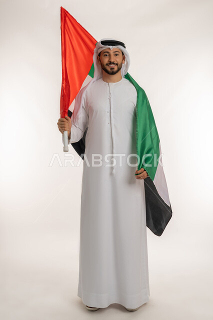 يوم استقلال الإمارات 1971 ، الوقوف باستقامة والنظر إلى الكاميرا بتعابير الفخر، بورتريه لرجل عربي خليجي إماراتي يرتدي الكندورة والغترة يحمل علم الإمارات العربية المتحدة على كتفه، هوية اليوم الوطني الاماراتي 2 ديسمبر، الطول الكامل للجسم، خلفية بيضاء