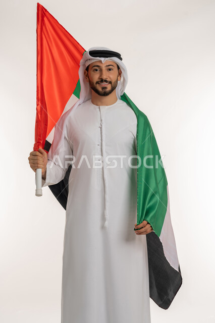 هوية اليوم الوطني الاماراتي 2  ديسمبر ، يوم استقلال الإمارات 1971، النظر إلى الكاميرا بتعابير الفخر، بورتريه لرجل عربي خليجي إماراتي يرتدي الكندورة والغترة يحمل علم الإمارات العربية المتحدة على كتفه، الاعتزاز بإنجازات البلاد، خلفية بيضاء