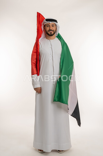 النظر إلى الكاميرا بتعابير الفخر، بورتريه لرجل عربي خليجي إماراتي يرتدي الكندورة والغترة يحمل علم الإمارات العربية المتحدة على كتفه، الاعتزاز بإنجازات البلاد، هوية اليوم الوطني الاماراتي 2  ديسمبر ، يوم استقلال الإمارات 1971 ، الطول الكامل للجسم، خلفية بيضاء