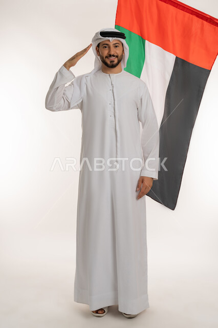 الأعياد والمناسبات الوطنية، ذكرى يوم الاستقلال الإماراتي 1971 ، بورتريه لرجل عربي خليجي إماراتي يرتدي الكندورة والغترة ينظر إلى الكاميرا وخلفه علم الإمارات، القاء التحية والاحترام للعلم، الاحتفال بيوم العلم الإماراتي 2 ديسمبر، الطول الكامل للجسم، خلفية بيضاء