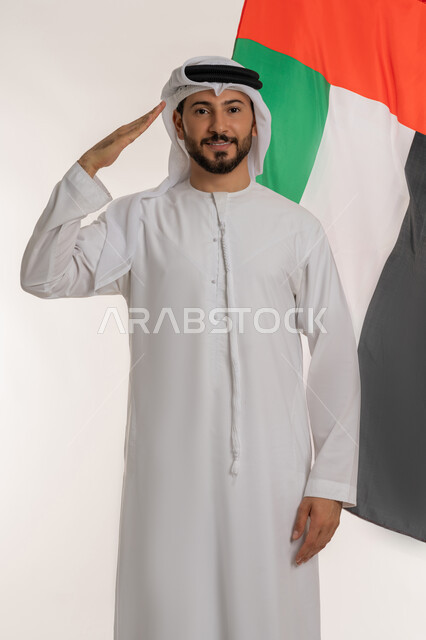 القاء التحية والاحترام للعلم الإماراتي، الاحتفال بيوم العلم الإماراتي 1971، الأعياد والمناسبات الوطنية، ذكرى يوم الاستقلال الإماراتي 2 ديسمبر، بورتريه لرجل عربي خليجي إماراتي يرتدي الكندورة والغترة ينظر إلى الكاميرا وخلفه علم دولة الإمارات، خلفية بيضاء