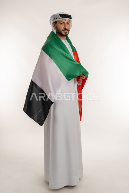 الحب والانتماء للوطن، النظر الى الكاميرا بايماءات الاعتزاز، هوية اليوم الوطني الاماراتي 2  ديسمبر، احياء ذكرى استقلال الإمارات ١٩٧١، بورتريه من الجانب لرجل عربي خليجي إماراتي يرتدي الكندورة والغترة يحمل علم الإمارات على ظهره، الطول الكامل للجسم، خلفية بيضاء