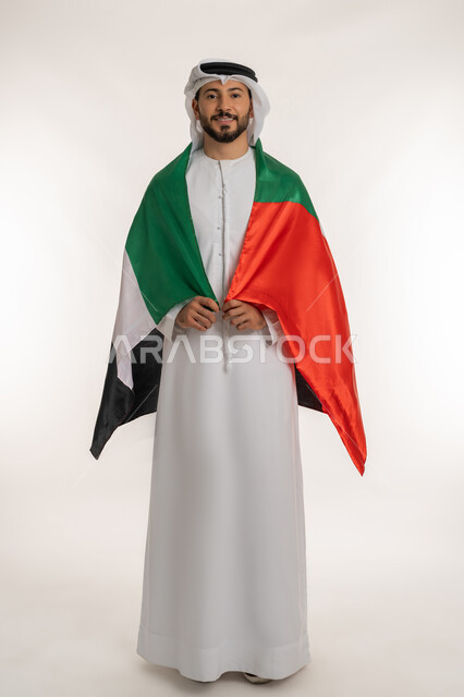 هوية اليوم الوطني الاماراتي 2 ديمسبر، احياء ذكرى استقلال الإمارات 1971 ، بورتريه لرجل عربي خليجي إماراتي يرتدي الكندورة والغترة يحمل علم الإمارات العربية المتحدة على ظهره، النظر الى الكاميرا بايماءات الاعتزاز والثقة، الطول الكامل للجسم، خلفية بيضاء
