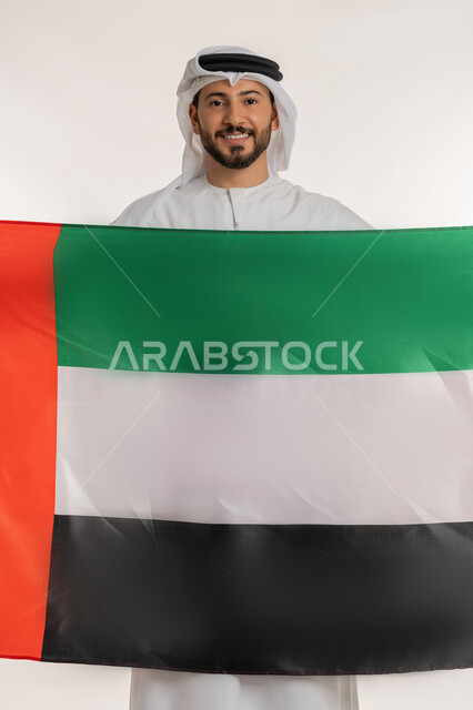 ذكرى استقلال الإمارات 1971، النظر الى الكاميرا بايماءات الاعتزاز والثقة، هوية اليوم الوطني الاماراتي 2 ديسمبر، بورتريه لرجل عربي خليجي إماراتي مبتسم يرتدي الكندورة والغترة يقوم يحمل علم الإمارات بتعابير المحبة والفخر، الحب والانتماء للوطن، خلفية بيضاء