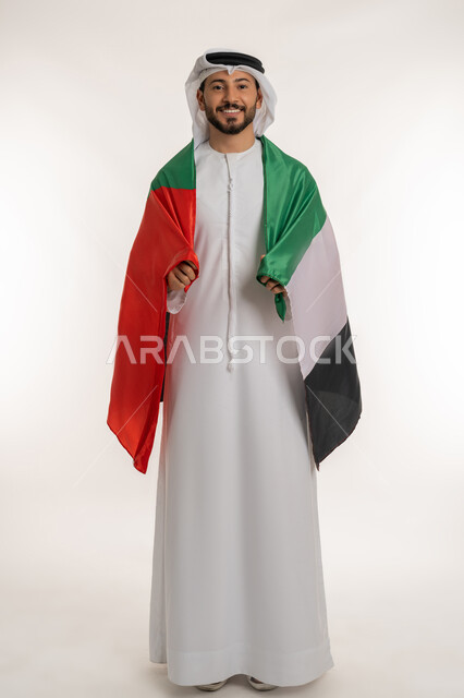 النظر الى الكاميرا بايماءات الاعتزاز والثقة، هوية اليوم الوطني الاماراتي 2 ديمسبر، احياء ذكرى استقلال الإمارات 1971 ، بورتريه لرجل عربي خليجي إماراتي يرتدي الكندورة والغترة يحمل علم الإمارات العربية المتحدة على ظهره، الطول الكامل للجسم، خلفية بيضاء
