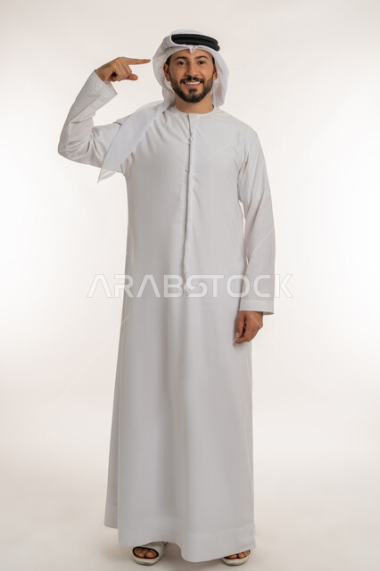 التفكير والتحليل و الذكاء والفطنه، بورتريه لرجل عربي خليجي إماراتي مبتسم يرتدي الكندورة والغترة ينظر الى الكاميرا يشير إصبعه السبابة الىرأسه بإيماءات البشاشة والسرور، مفهوم الإصرار على الرأي وفعل مايرغب، الطول الكامل للجسم, خلفية بيضاء