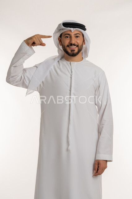 مفهوم الإصرار على الرأي وفعل مايرغب، التفكير والتحليل و الذكاء والفطنه، بورتريه لرجل عربي خليجي إماراتي مبتسم يرتدي الكندورة والغترة ينظر الى الكاميرا يشير إصبعه السبابة الىرأسه بإيماءات البشاشة والسرور، خلفية بيضاء