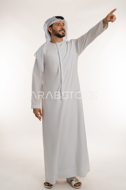 تحديد موقع معين بتعابير البشاشة, الاشارة بإصبع السبابة إلى هدف محدد, الدلالة على مكان عنصر, بورتريه لرجل عربي خليجي إماراتي مبتسم يرتدي الكندورة والغتره ينظر إلى شيء ما بإيماءات السعادة و السرور، خلفية بيضاء