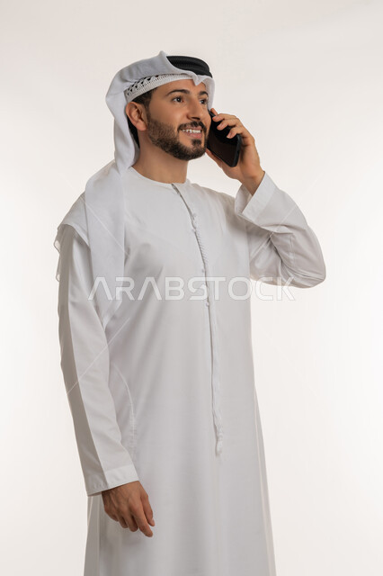 التواصل مع الأهل والأصدقاء، إيماءات السعادة والسرور لسماع الأخبار المفرحة، بورتريه لرجل عربي خليجي إماراتي مبتسم يرتدي الكندورة والغترة يقوم بإجراء مكالمة هاتفية عبر الهاتف المحمول، استخدام التقنيات والأجهزة الحديثة، خلفية بيضاء