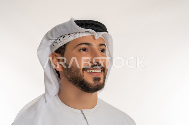 الوقوف باستقامة واعتدال، مفهوم الرجولة والثقة بالنفس، الاهتمام بالأناقة والمظهر الخارجي، بورتريه مقرب لرجل عربي خليجي إماراتي مبتسم يرتدي الكندورة والغترة ينظر الى شيء ما، ايماءات السعادة والسرور، خلفية بيضاء