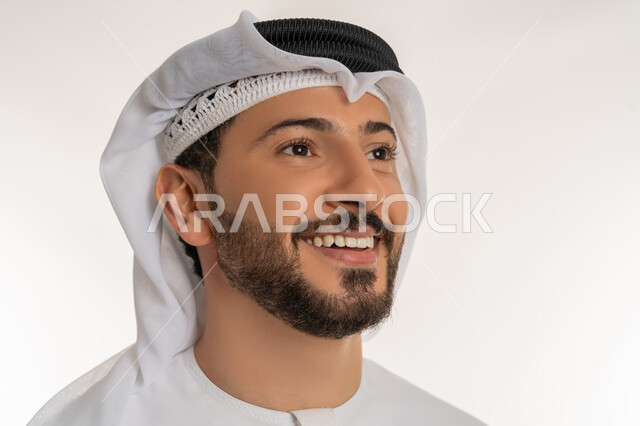 ايماءات السعادة والسرور، الوقوف باستقامة واعتدال، مفهوم الرجولة والثقة بالنفس، الاهتمام بالأناقة والمظهر الخارجي، بورتريه مقرب لرجل عربي خليجي إماراتي مبتسم يرتدي الكندورة والغترة ينظر الى شيء ما، خلفية بيضاء