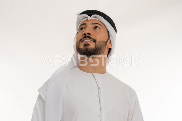 Concept of masculinity and self-confidence, interest in elegance and appearance, close-up portrait of a smiling foreign man wearing a kandura and ghutra looking at something, gestures of happiness and pleasure, taking a selfie for official documents, standing straight and upright, white background