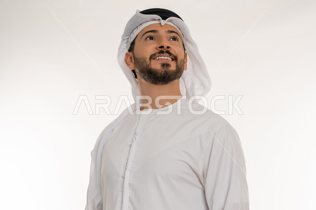 الوقوف باستقامة واعتدال، مفهوم الرجولة والثقة بالنفس، الاهتمام بالأناقة والمظهر الخارجي، بورتريه مقرب لرجل عربي خليجي إماراتي مبتسم يرتدي الكندورة والغترة ينظر الى شيء ما، ايماءات السعادة والسرور، خلفية بيضاء