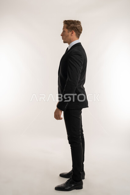 الاهتمام بالمظهر الخارجي، مفهوم الأناقة والرجولة والقوة, بورتريه من الجانب لرجل أعمال أجنبي يرتدي البدلة الرسمية ينظر الى شيء ما, الوقوف باستقامة وثبات والنظر الى اليسار بايماءات الثقة بالنفس, تعابير الجدية والحزم، الطول الكامل للجسم، خلفية بيضاء