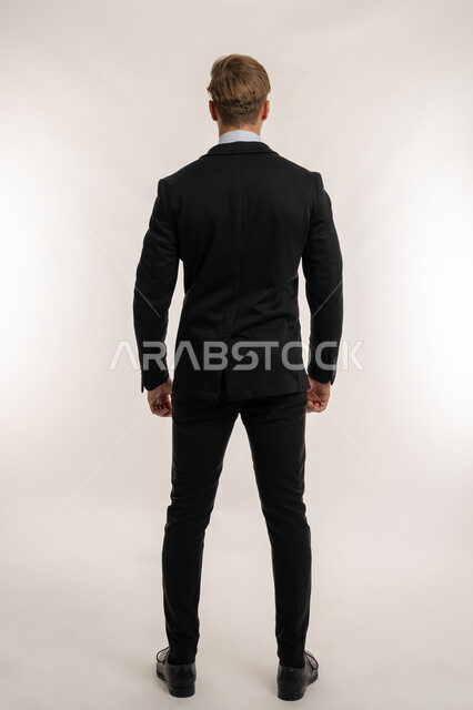 مفهوم الرجولة والقوة، الأناقة والاهتمام بالمظهر الخارجي، بورتريه من الخلف لرجل أعمال أجنبي يرتدي البدلة الرسمية ينظر إلى شي ما، الوقوف باستقامة وثبات، الاعتراض عن موضوع محدد، الطول الكامل للجسم، خلفية بيضاء