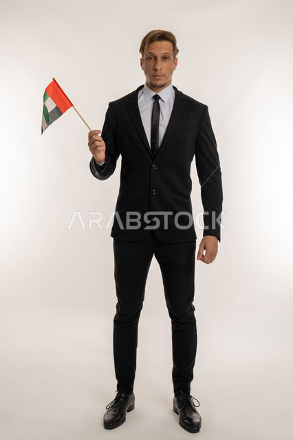 التعرف على ثقافات وتاريخ الامارات، بورتريه لرجل اعمال اجنبي مبتسم يرتدي البدلة الرسمية يحمل العلم للاحتفال باليوم بالوطني الاماراتي 2 ديسمبر، الوقوف باستقامة والنظر إلى الكاميرا بتعابير الفخر والاعتزاز، ذكرى تأسيس الاتحاد 1971، الطول الكامل للجسم، خلفية بيضاء