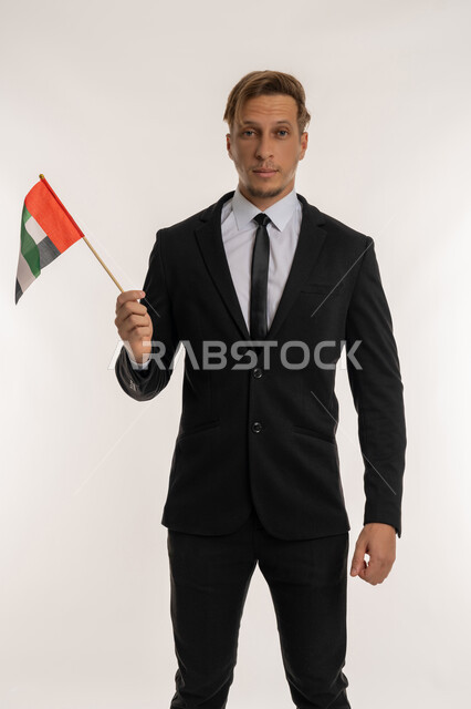 Union Day 1971, Getting to know the cultures and history of the Emirates, Portrait of a foreign businessman wearing a formal suit holding a flag to celebrate the UAE National Day, December 2, standing straight and firm with expressions of pride and honor, looking at the camera with gestures of pleasure, white background