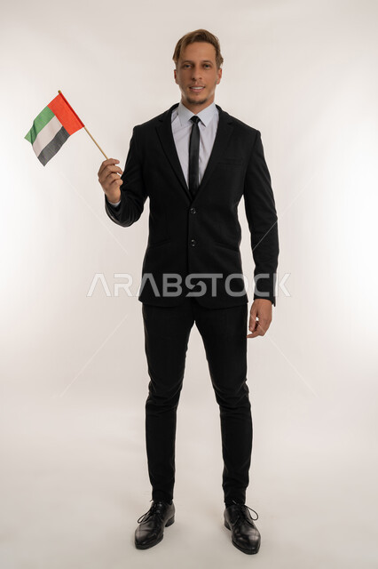 Getting to know the culture and history of the Emirates, Portrait of a foreign businessman wearing a formal suit holding a flag to celebrate the UAE National Day, December 2, standing straight and looking at the camera with expressions of pride and honor, Anniversary of the founding of the Union 1971, full body length, white background
