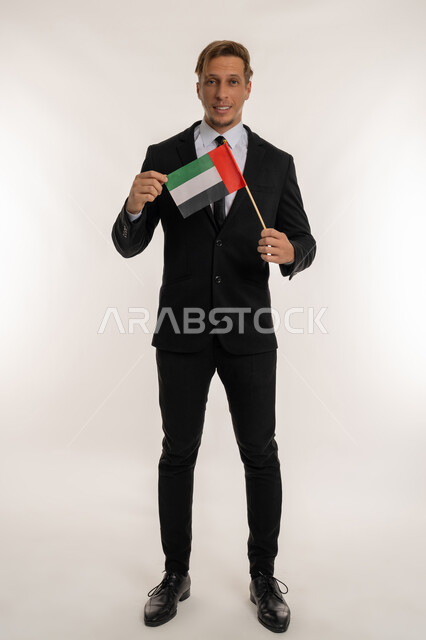 الحب والانتماء للوطن، التعرف على ثقافات وتاريخ الامارات، بورتريه لرجل اعمال اجنبي مبتسم يرتدي البدلة الرسمية يحمل العلم للاحتفال باليوم بالوطني الاماراتي 2 ديسمبر، النظر إلى الكاميرا بتعابير الفخر، ذكرى تأسيس الاتحاد 1971، الطول الكامل للجسم، خلفية بيضاء