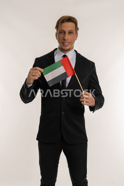 النظر إلى الكاميرا بايماءات السرور، ذكرى تأسيس الاتحاد 1971، التعرف على ثقافات وتاريخ الامارات، بورتريه لرجل اعمال اجنبي مبتسم يرتدي البدلة الرسمية يحمل العلم للاحتفال باليوم بالوطني الاماراتي 2 ديسمبر، الوقوف باستقامة وثبات بتعابير الفخر والاعتزاز، خلفية بيضاء